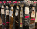 Six packaged bundles of hair extensions displayed on a black pegboard rack. The bundles are in different shades including brown, blonde, and black, each wrapped in clear plastic with a white brand label and a red size/variant header card at the top.