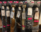 Six packaged bundles of hair extensions displayed on a black pegboard rack. The bundles are in different shades including brown, blonde, and black, each wrapped in clear plastic with a white brand label and a red size/variant header card at the top.