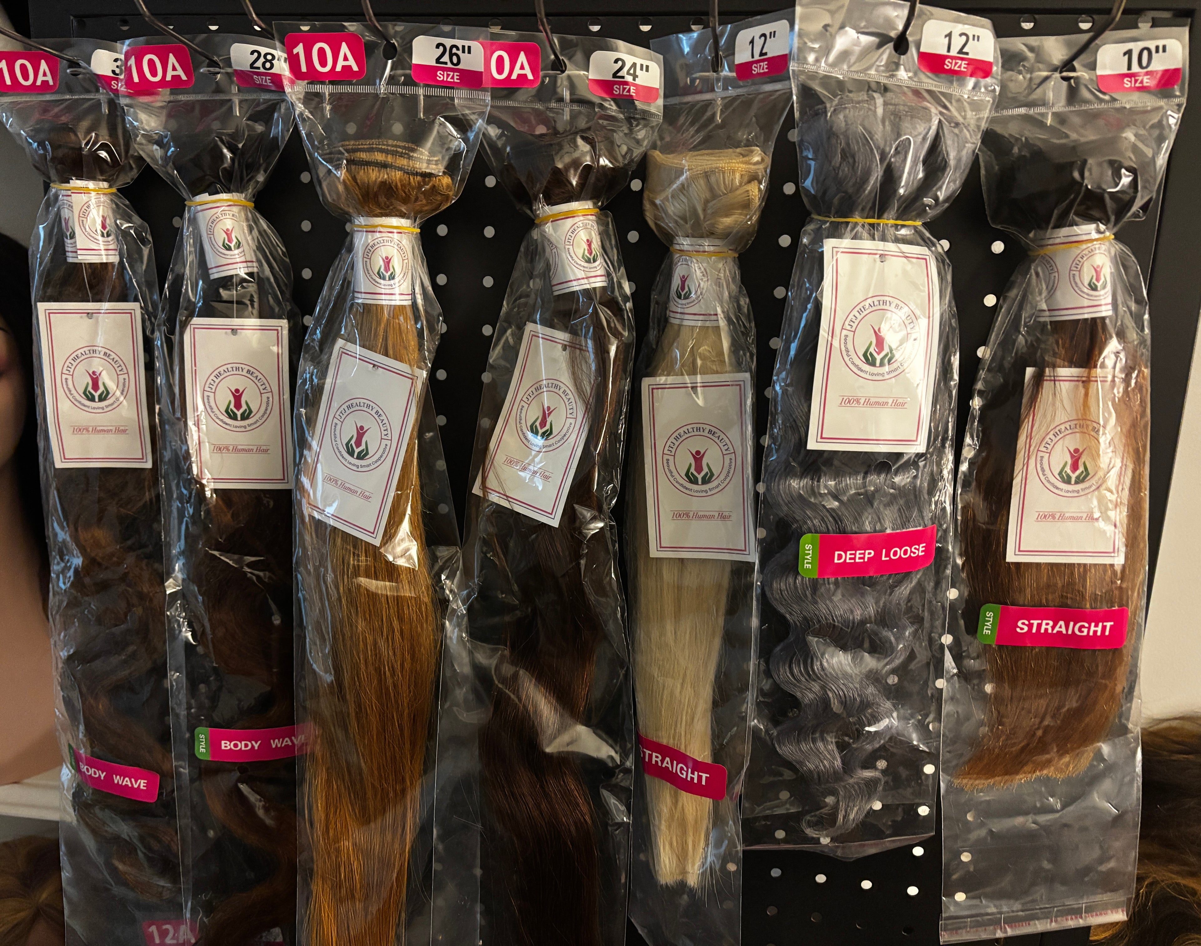 Six packaged bundles of hair extensions displayed on a black pegboard rack. The bundles are in different shades including brown, blonde, and black, each wrapped in clear plastic with a white brand label and a red size/variant header card at the top.