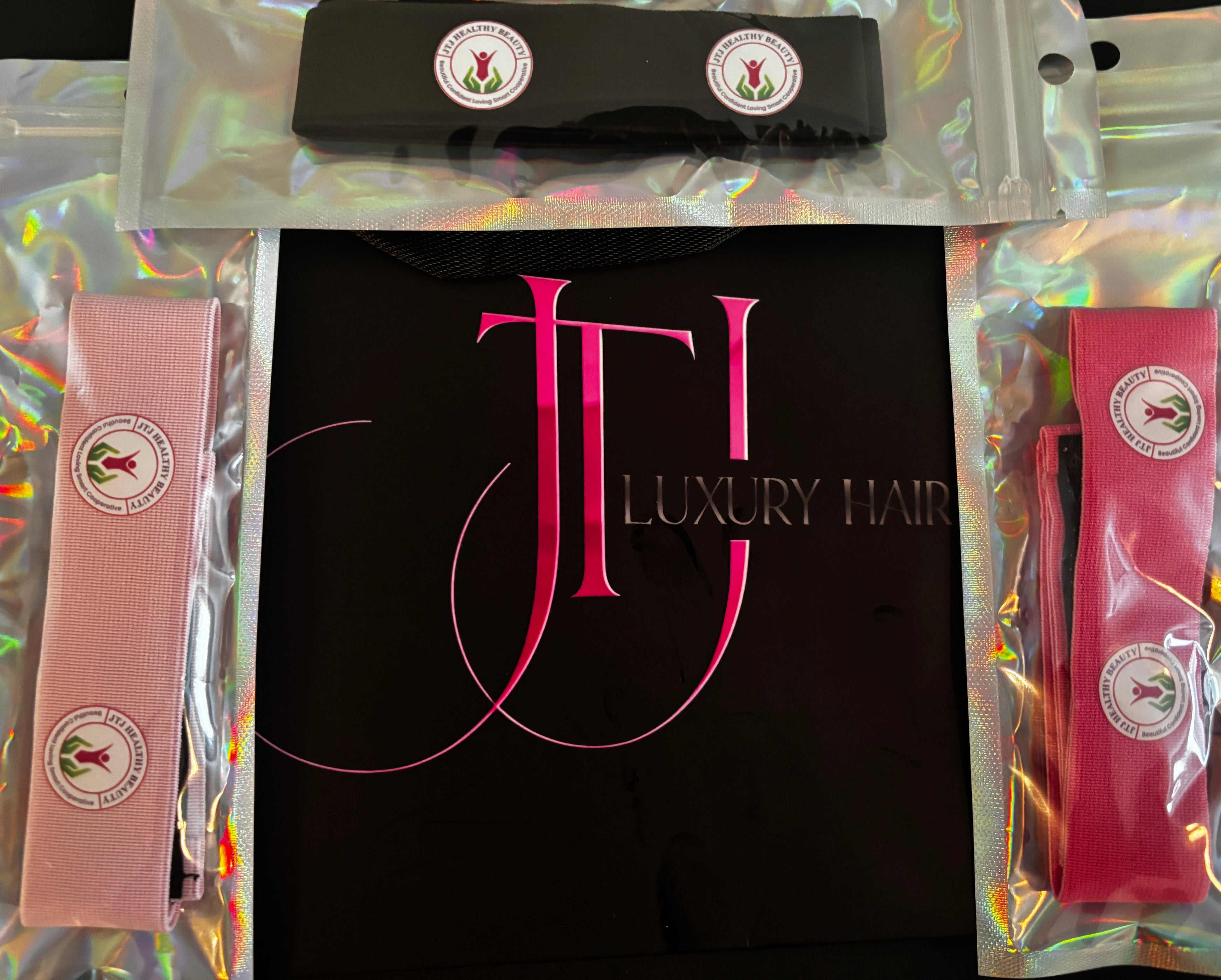 A luxury hair accessory set featuring pink and black headbands with circular logo patches, packaged in clear sleeves with holographic backing, alongside a black branded box.