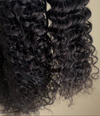 Two bundles of dark brown to black curly wig hair in a clear plastic bag, with a small pink and white tag visible on the left, against a plain beige background.